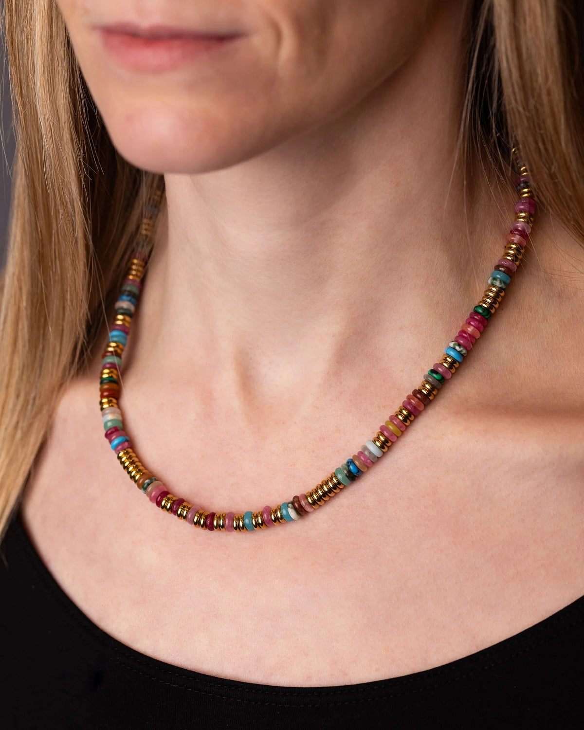 A close-up of a woman wearing a colourful beaded necklace with small metallic and painted disc-like beads in pink, blue, gold, and green tones.