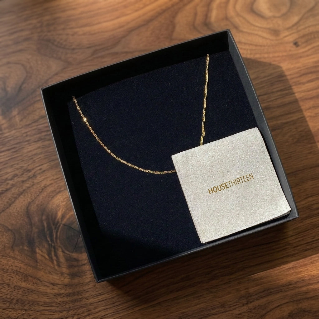 A delicate gold Twist chain necklace presented in an open black jewellery box with a black velvet insert and a branded light grey pouch in the bottom right corner. The card reads HOUSE THREEN and is placed on a wooden tabletop.