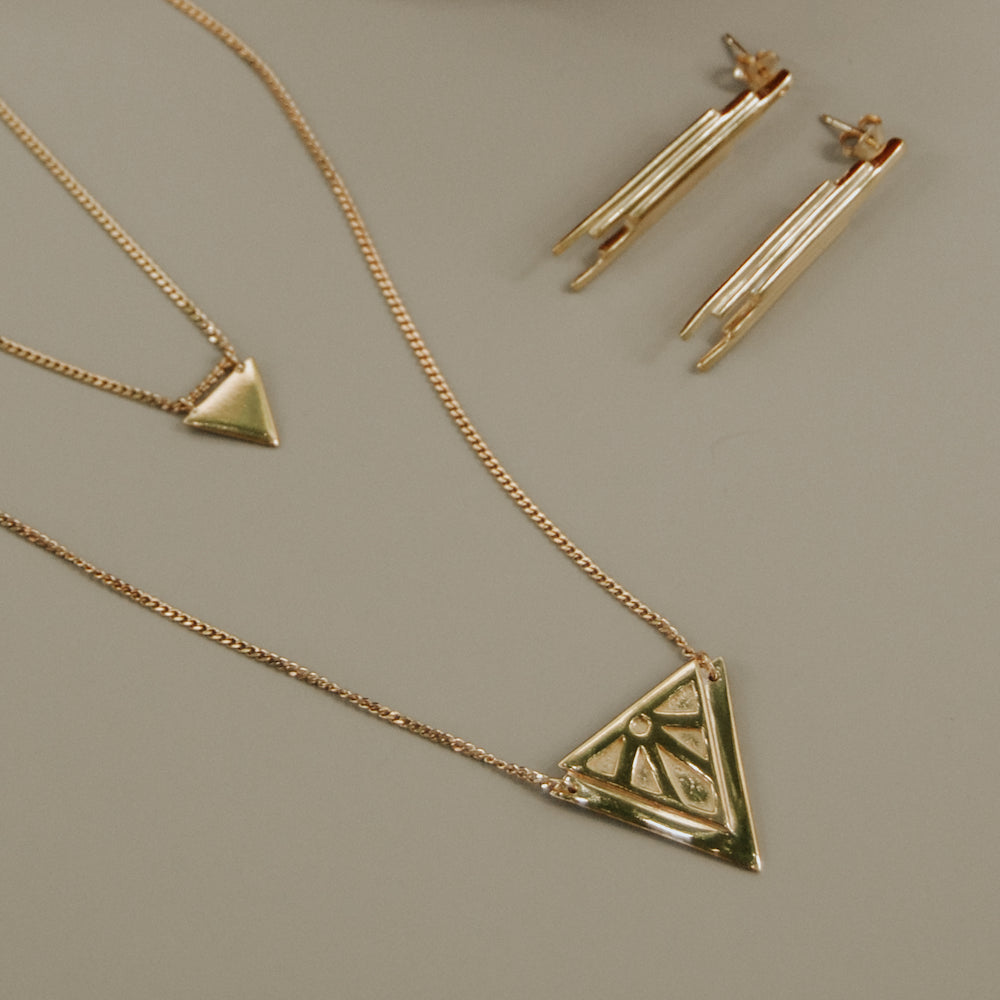 Gold Rise jewellery set featuring a triangle pendant necklace, a smaller matching triangle pendant on a chain, and a pair of Stud earrings, arranged on a neutral taupe background.