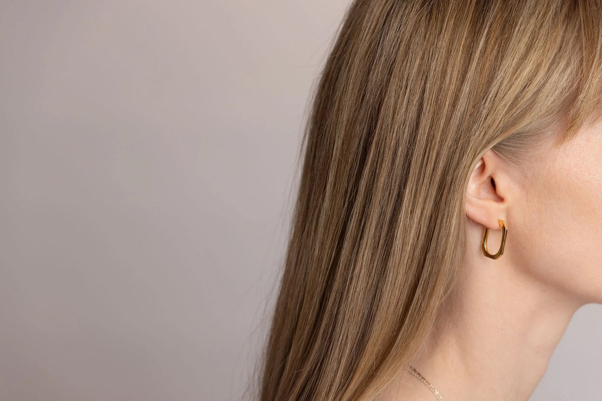 A close-up side profile of a woman with long blonde hair wearing a gold Square hoop earring against a plain light grey background.