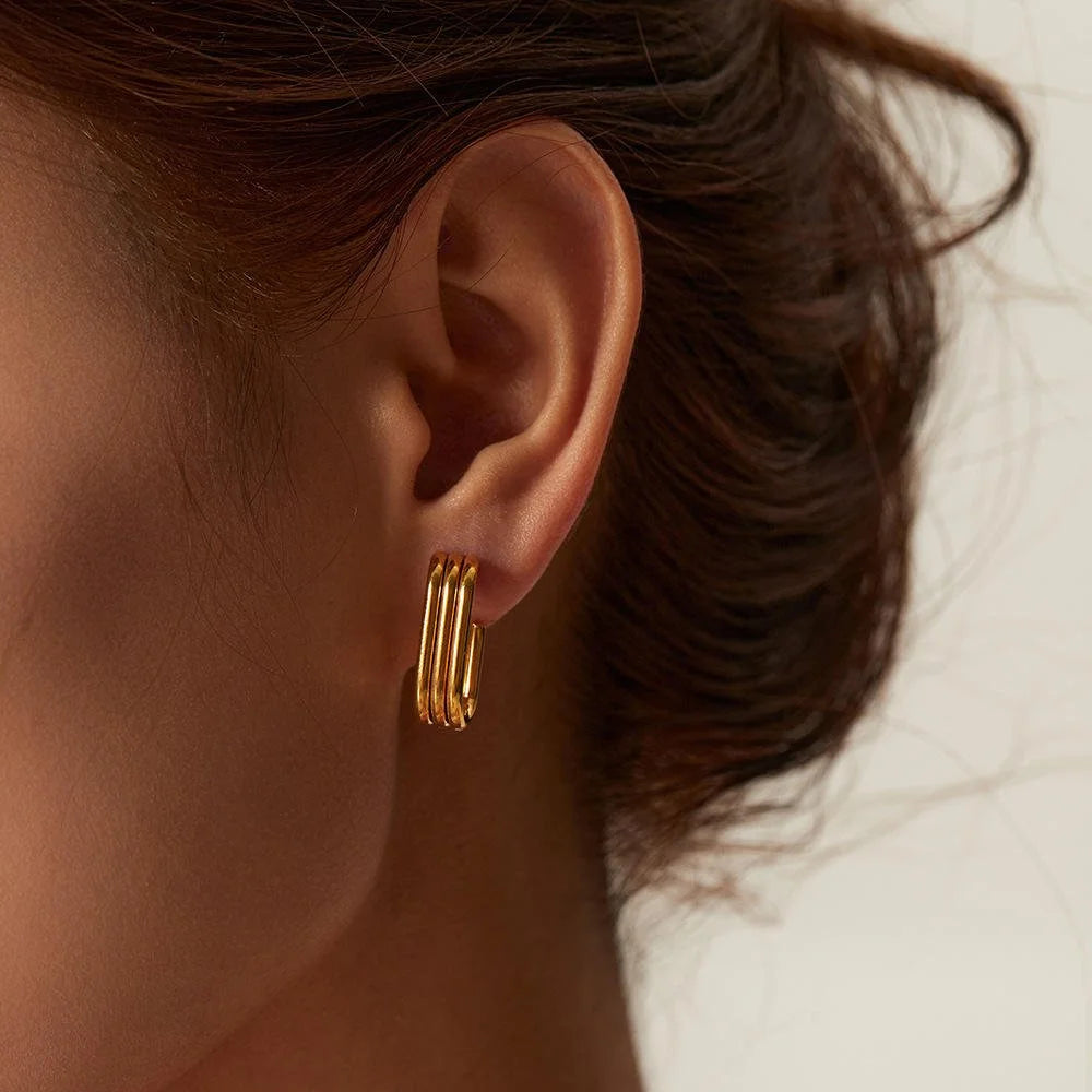 A close-up of a model's ear wearing a gold Trio hoop earring with three parallel wire bands forming a rectangular hoop, set against a soft, light neutral background.