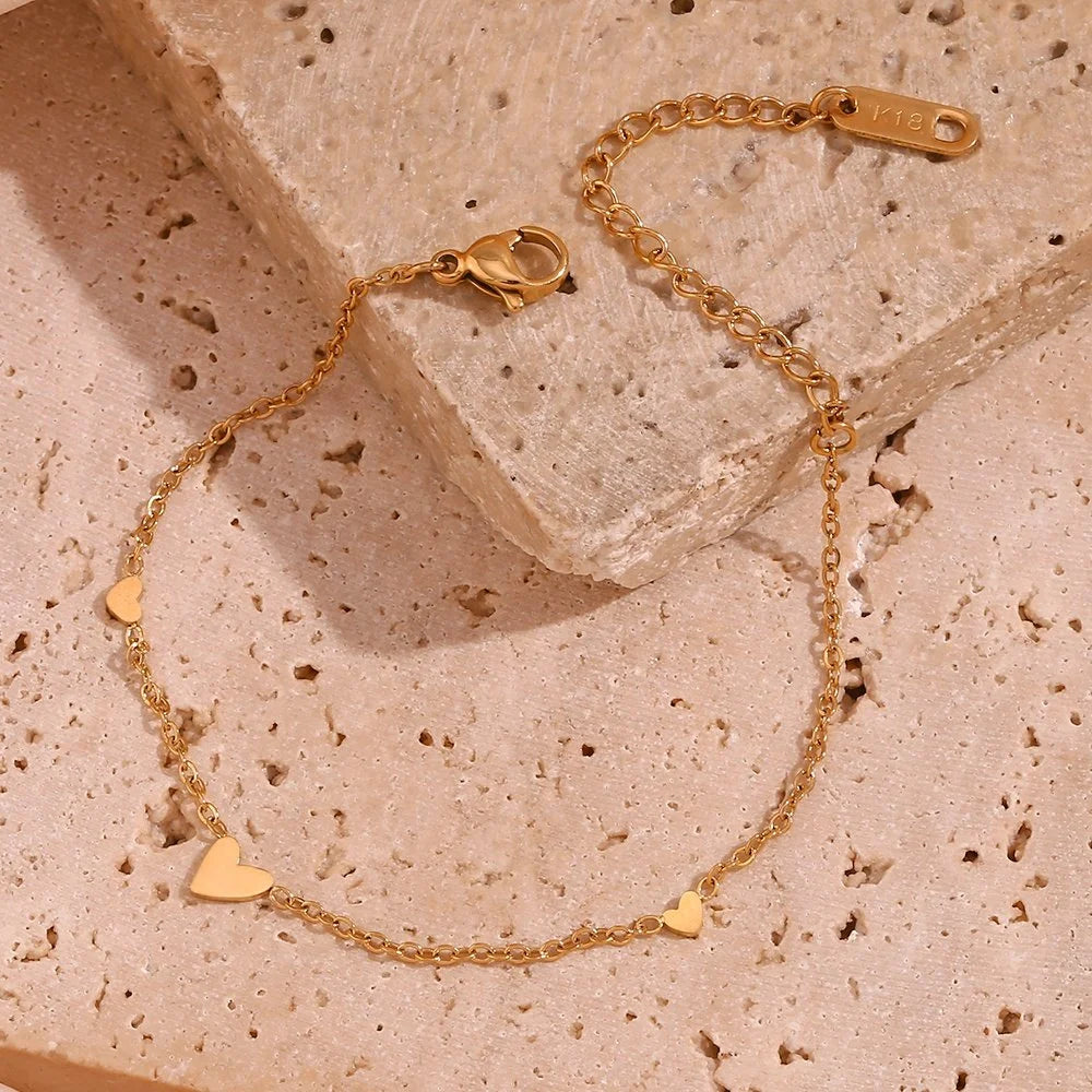 A delicate gold chain bracelet with three small heart charms, displayed on a textured beige stone surface.