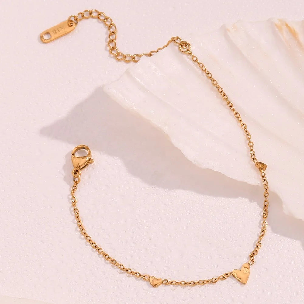 A delicate gold chain anklet with small heart charms, displayed on a textured white surface with a white shell prop.