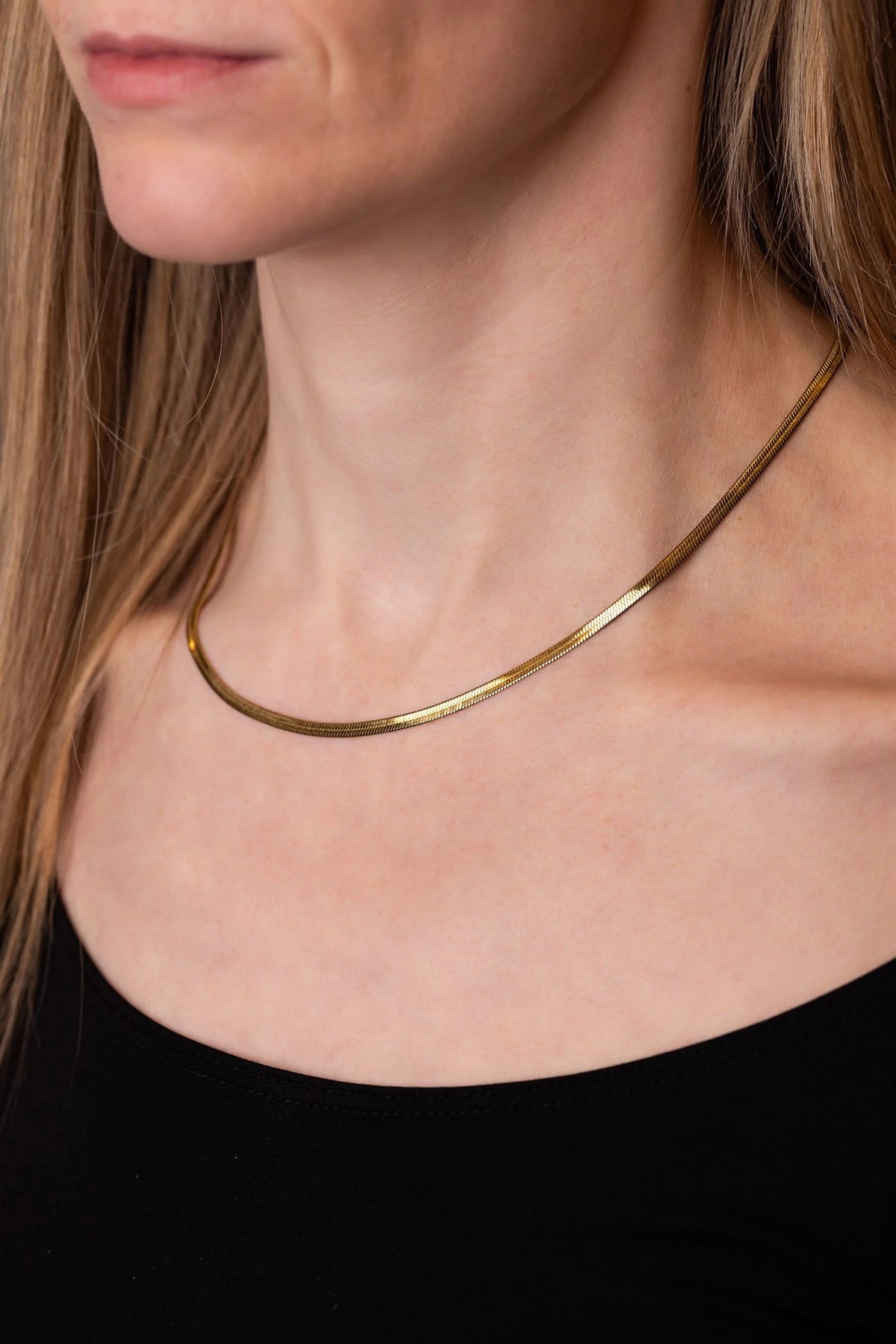 A close-up of a model wearing a thin, polished gold snake chain necklace against her neck and collarbone, paired with a black top.