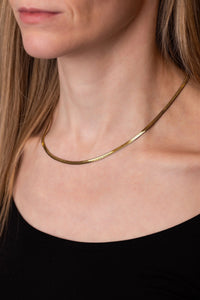 A close-up of a model wearing a thin, polished gold snake chain necklace against her neck and collarbone, paired with a black top.