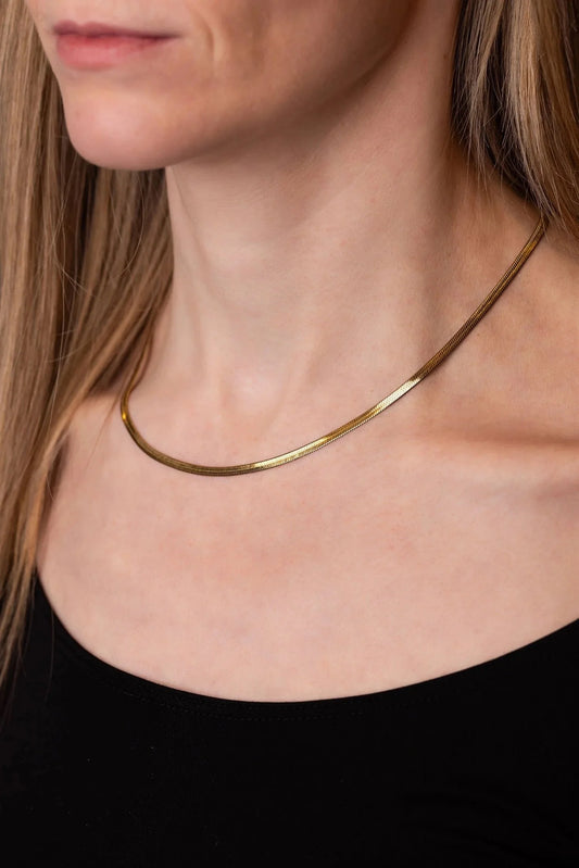 A close-up of a model wearing a thin, polished gold Herringbone chain necklace with a black top.