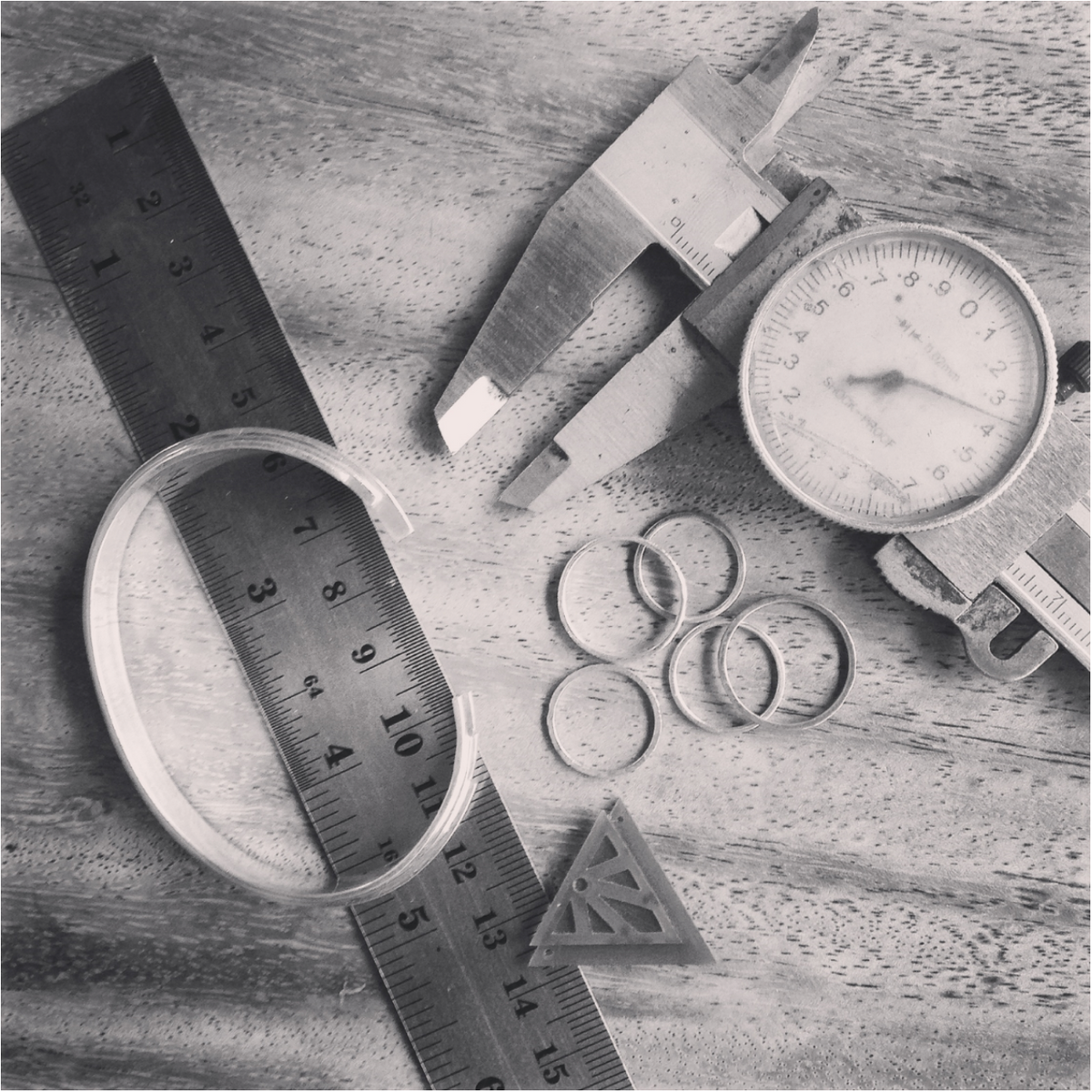 A black and white photograph showing jewellery measuring tools and rings arranged on a textured wooden surface, including a metal ruler, callipers, a dial gauge, several rings, and a triangular wax carving.