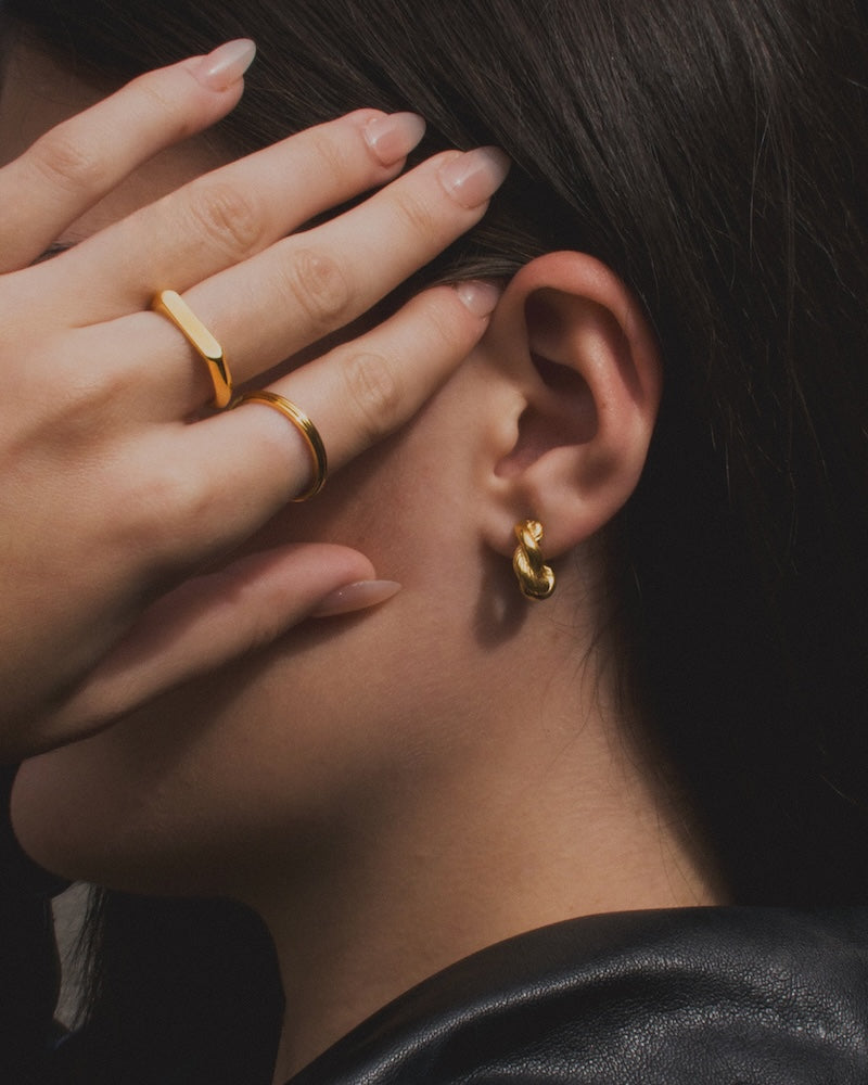 Woman with dark hair stood to the side with her hand covering the side of her face with two gold rings on her fingers. She also wears the medium sized twisted knot hoops