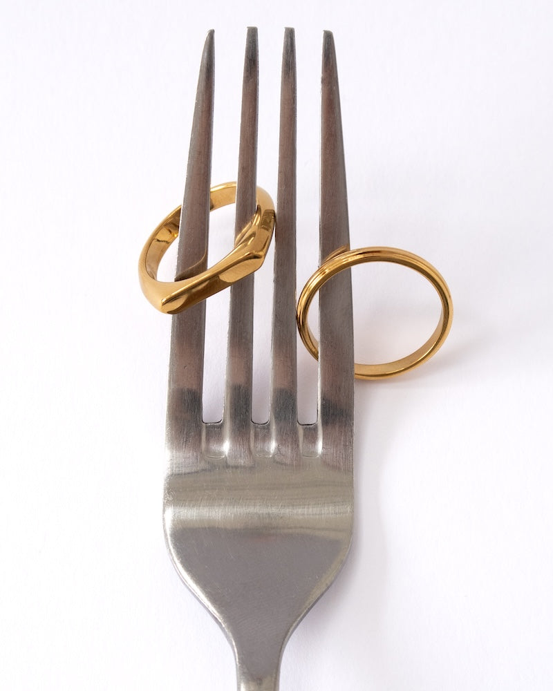 Two gold rings, including the Ridge Stacking Ring and Slim Signet Ring , displayed on the tines of a silver metal fork against a plain white background.