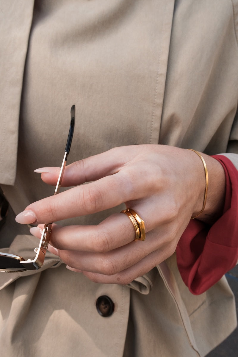 A close-up of a person's hand wearing two stacked gold rings, holding a pair of sunglasses, dressed in a beige trench coat and a red shirt.