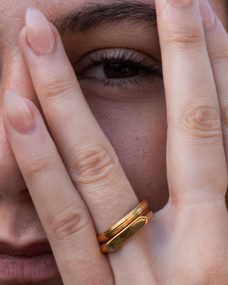 A close-up of a person's hand and eye area wearing a polished gold ring with a flat, elongated oval face and on one finger. second ridge stacking ring both on one finger. 