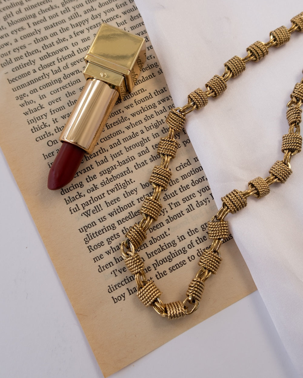 A gold-tone lipstick case with a deep red lipstick bullet and a chunky gold-tone chain necklace, styled on top of an open book page.