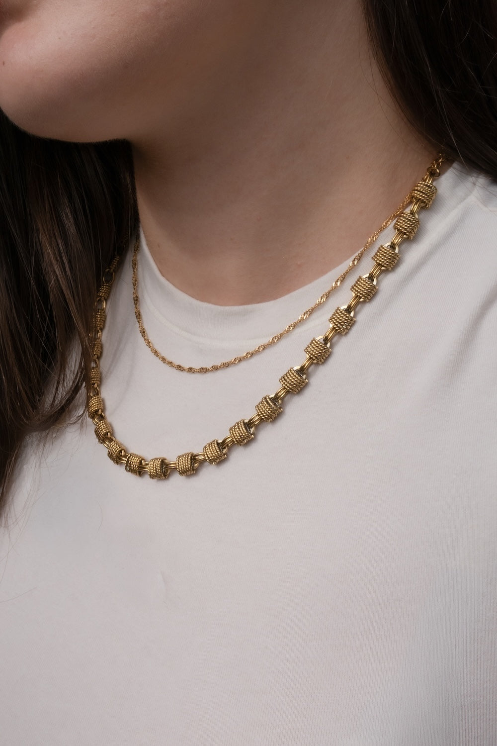 A close-up of a person wearing a textured gold Woven Chunky chain necklace over a thin gold Twist chain, paired with a white t-shirt.