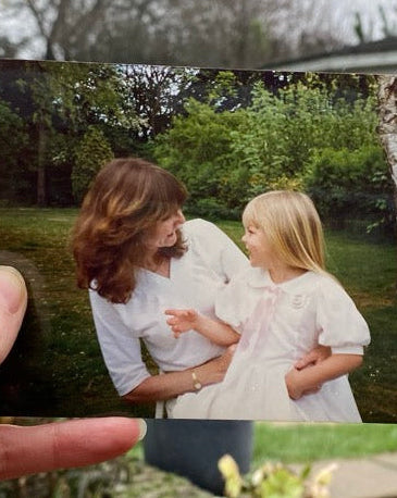 A hand holds a printed photograph in front of a real garden and house background. The photo shows a woman and a child in white clothing outdoors.