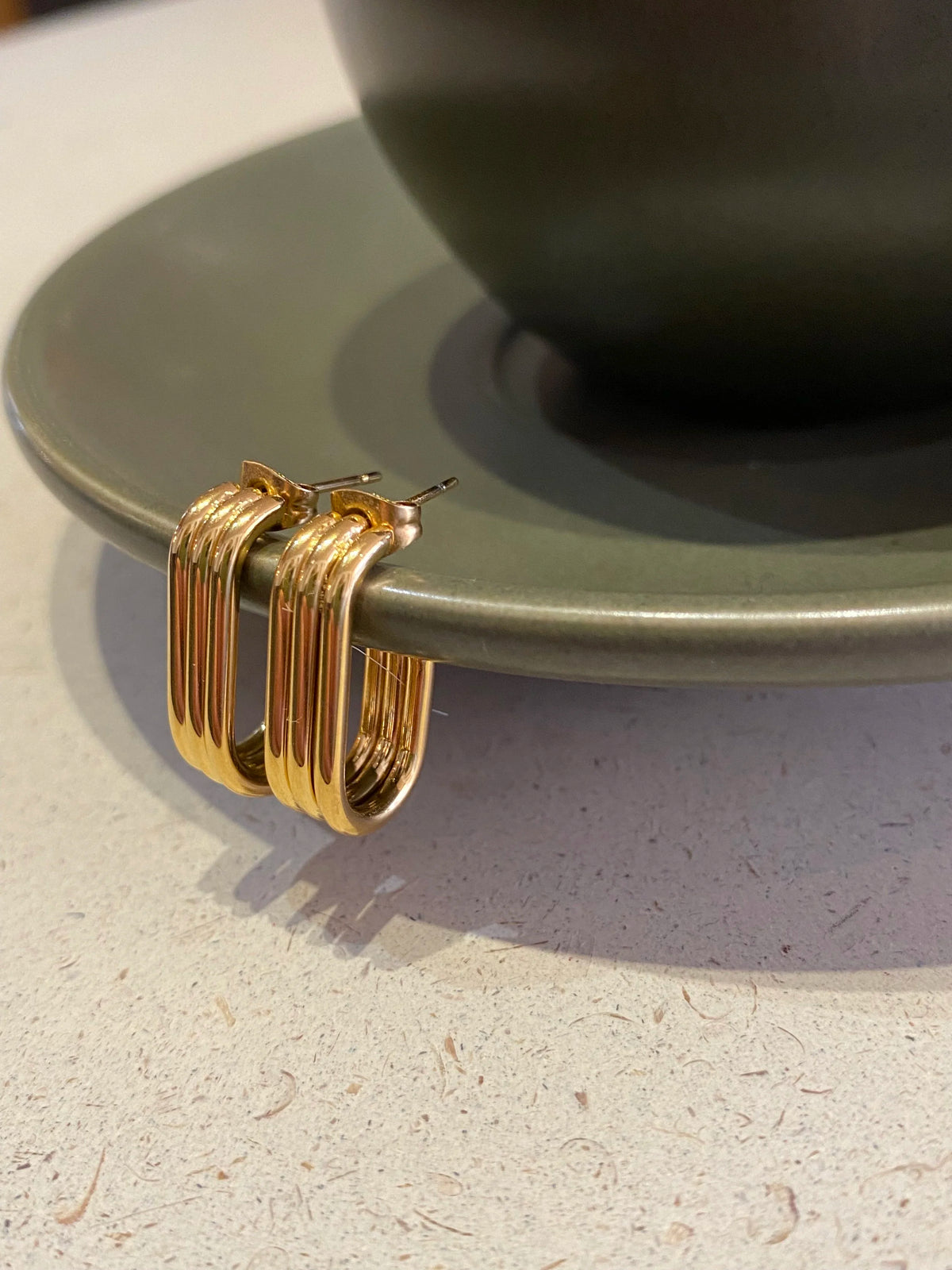 A pair of gold-toned Trio hoop earrings with three parallel vertical bands, hanging from a green ceramic saucer with a green cup on it, on a speckled light beige countertop.