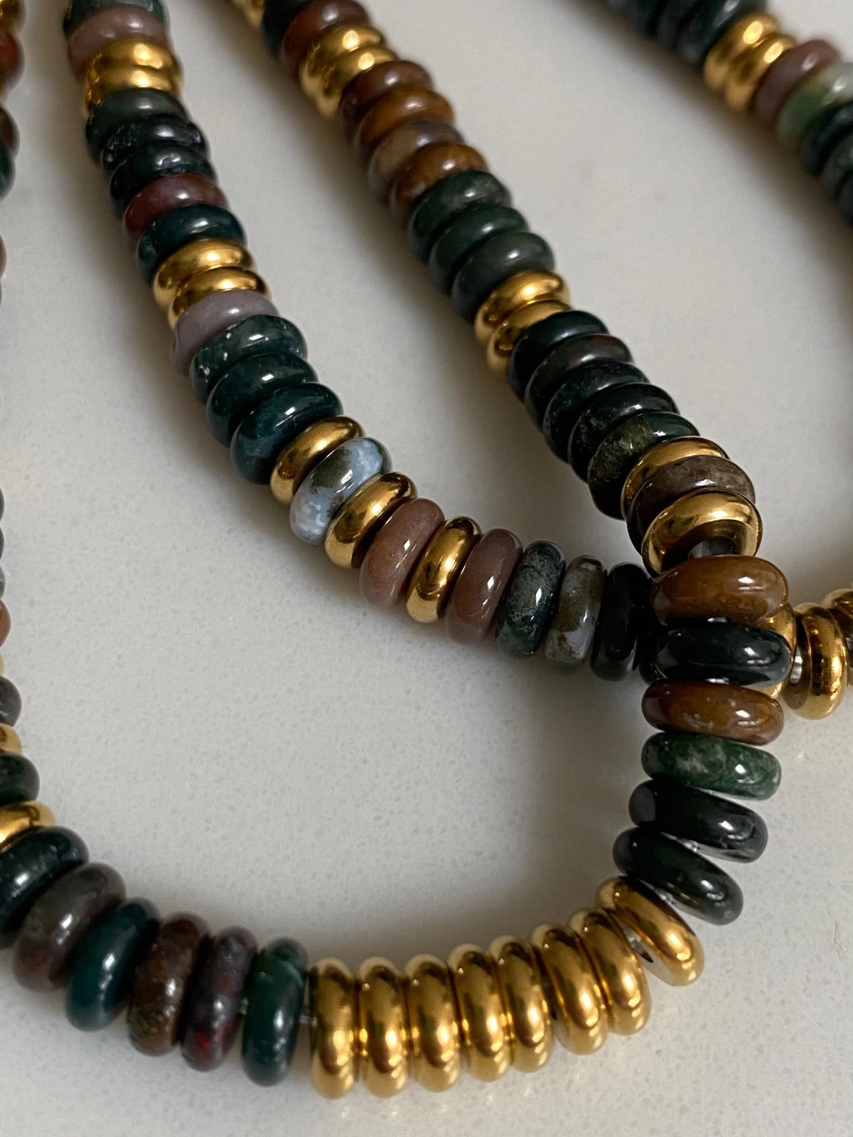 A close-up of a beaded Indian Agate featuring alternating polished dark green, brown, black and gold gemstones, laid out on a light neutral surface.