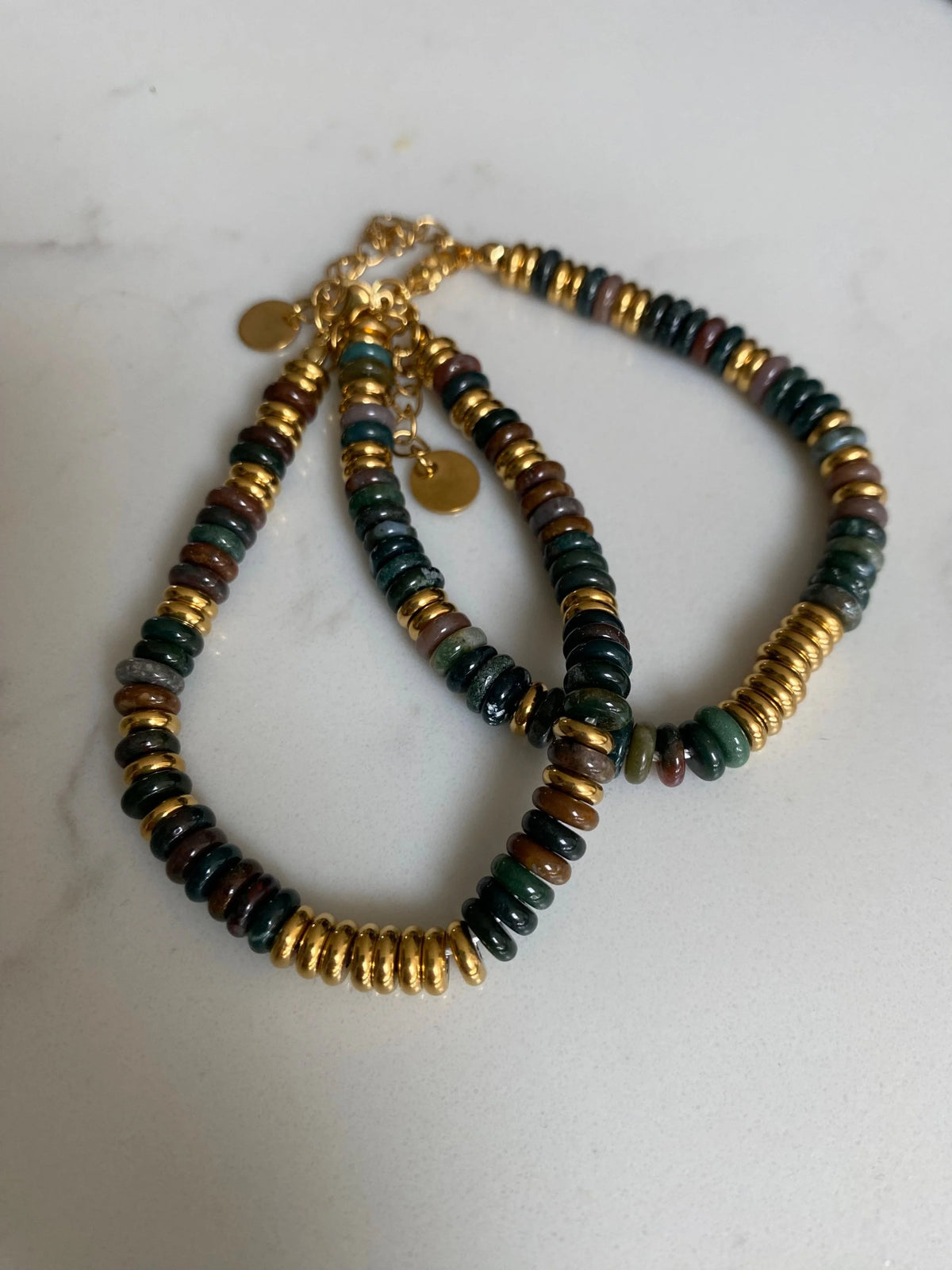 Indian agate  bracelet with alternating dark green, brown, and metallic gold disc beads, featuring a gold clasp with two small round tags, displayed on a light off-white marble surface.