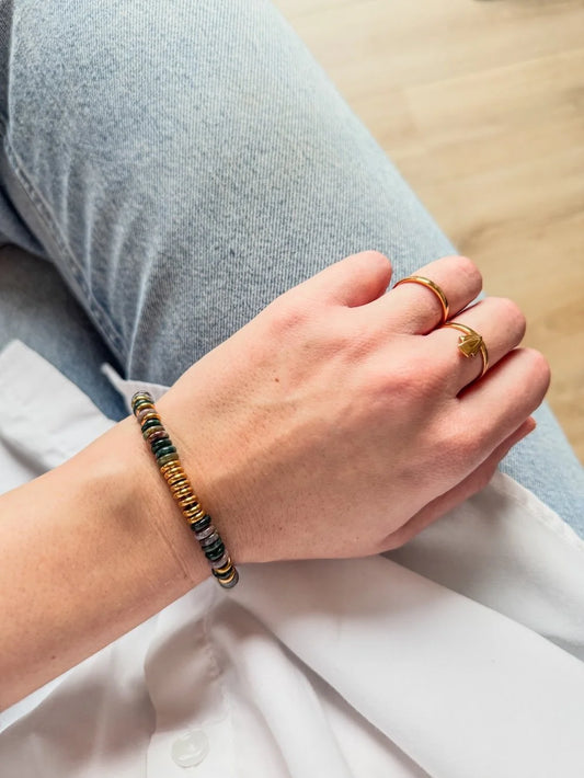 A close-up of a person's hand resting on their lap, wearing a beaded bracelet with gold, green, gold and black gemstones, along with gold rings, against a background of light blue jeans and a white shirt.
