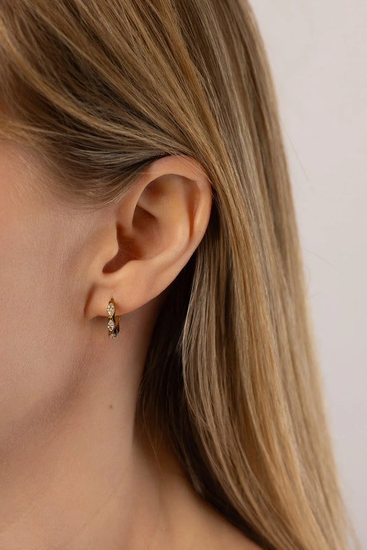 A close-up of a model's ear wearing a small gold Luna huggie hoop earring with tiny clear cubic zirconia crystals, against a plain light background.