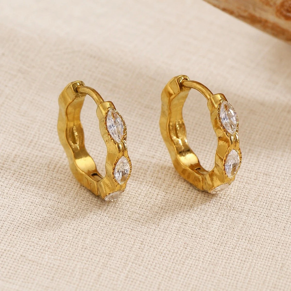 A pair of gold huggie hoop earrings with marquise-cut clear cubic zirconia crystals, displayed on a textured beige fabric background.