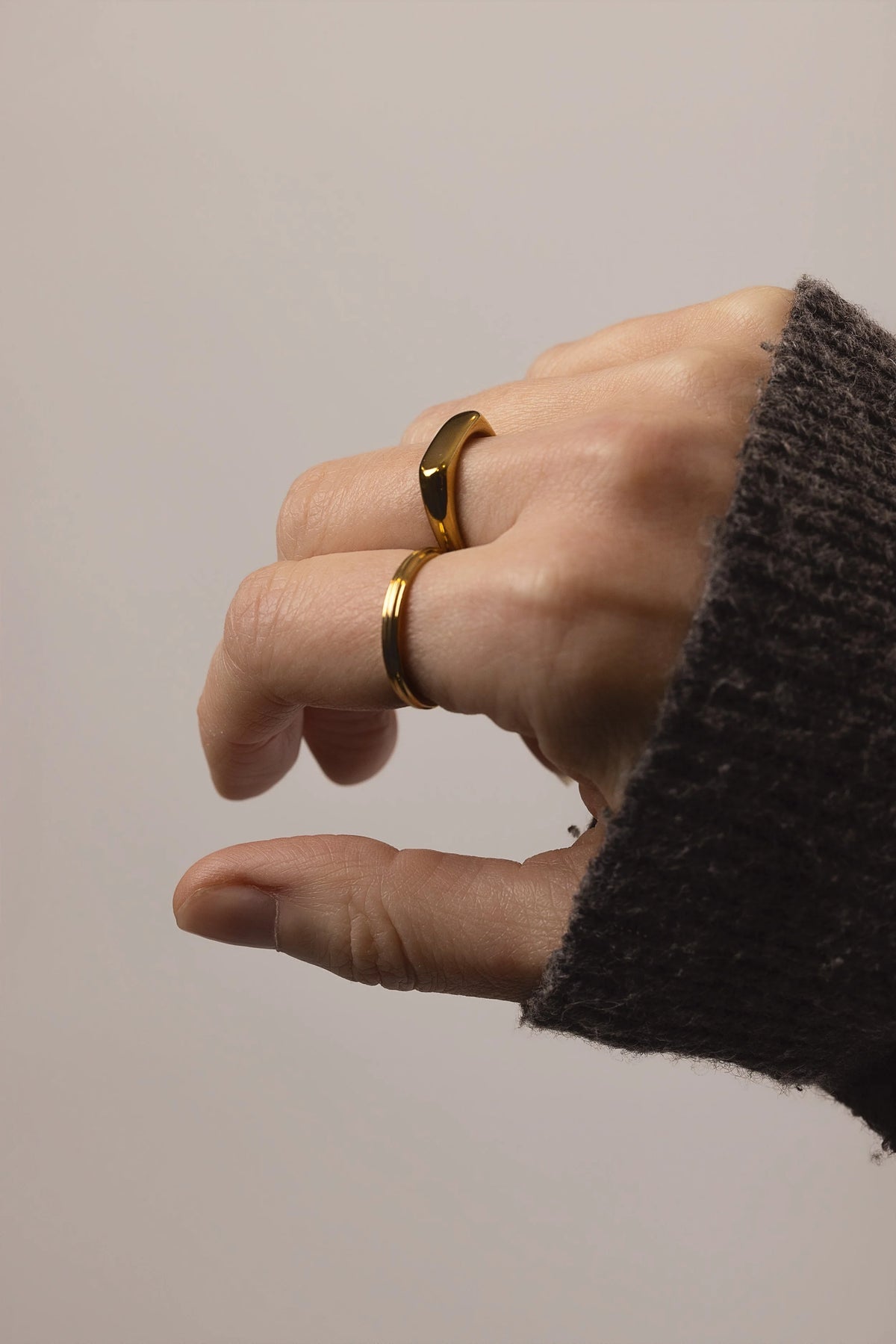 A close-up of a hand wearing two slim gold rings, one with a polished beveled edge (Slim Signet ring) and the other a Ridge stacking ring, set against a plain light grey background.