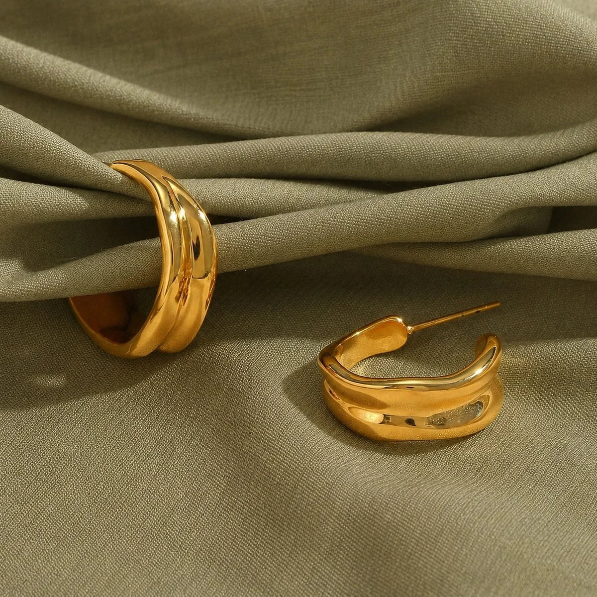 A pair of polished gold Molten hoop earrings with a molten texture displayed on draped olive green fabric.