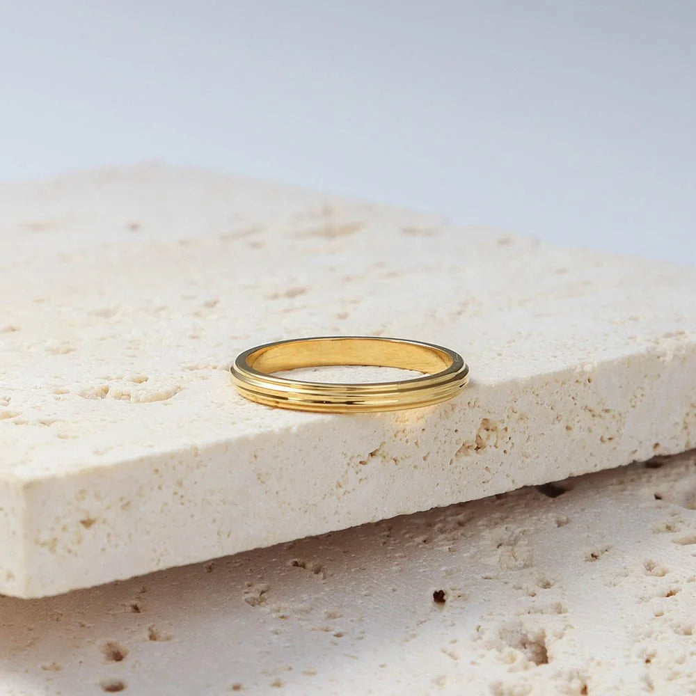 A thin gold band ring with a subtle ridged edge design, displayed on a textured beige stone slab against a light grey background.