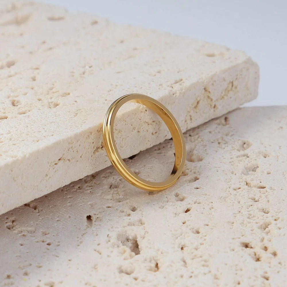 A minimalist gold band ring resting on a textured beige stone slab against a light gray background.