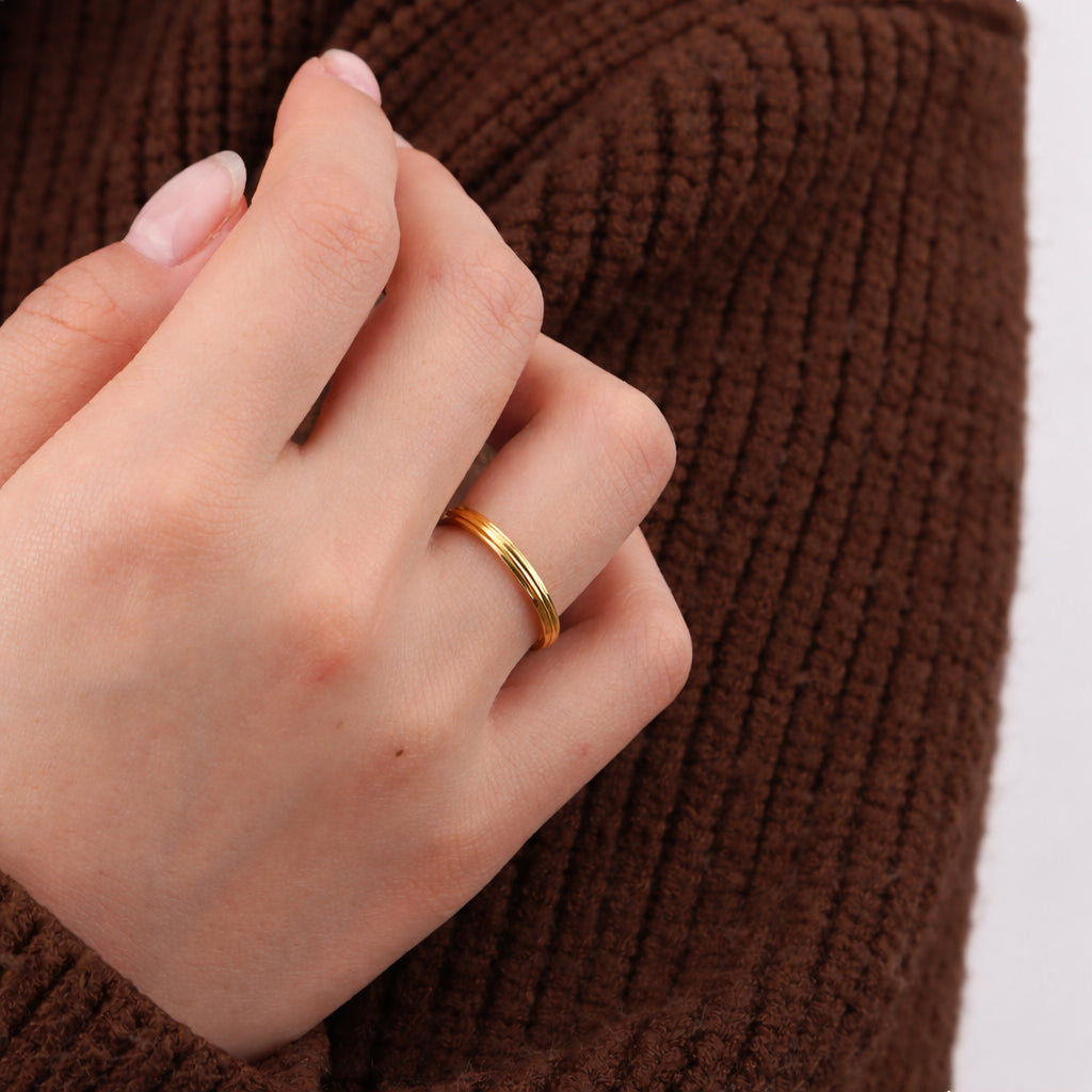 A close-up of a person's hand wearing a thin, polished gold band ring, set against a brown ribbed knit sweater.