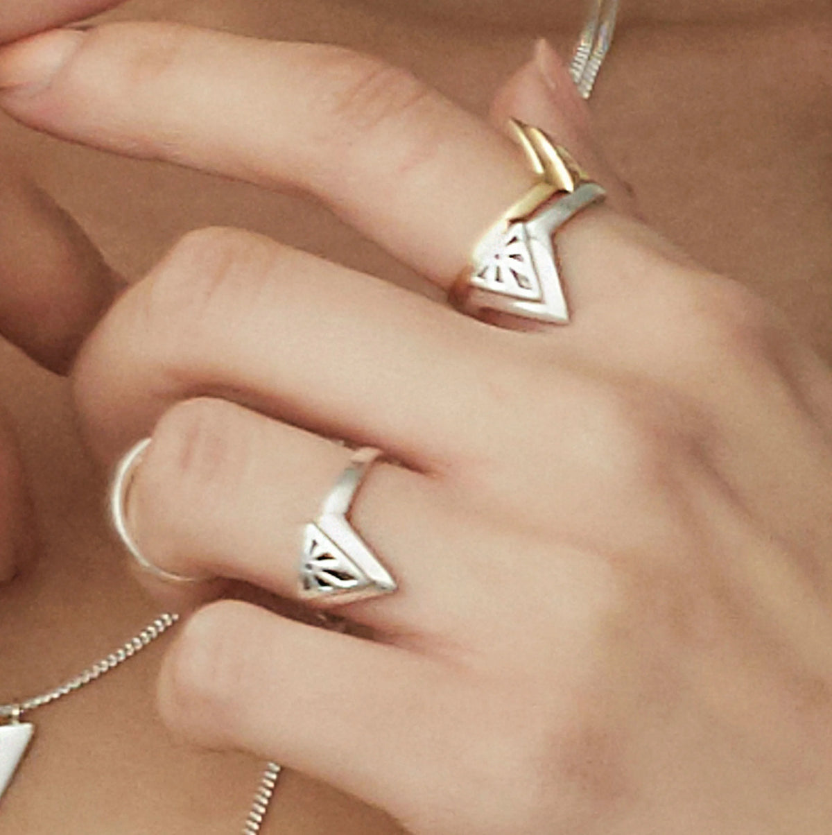 A close-up of a hand wearing multiple silver rings, including two triangular Rise rings and a gold Rise ring, with a silver chain visible.
