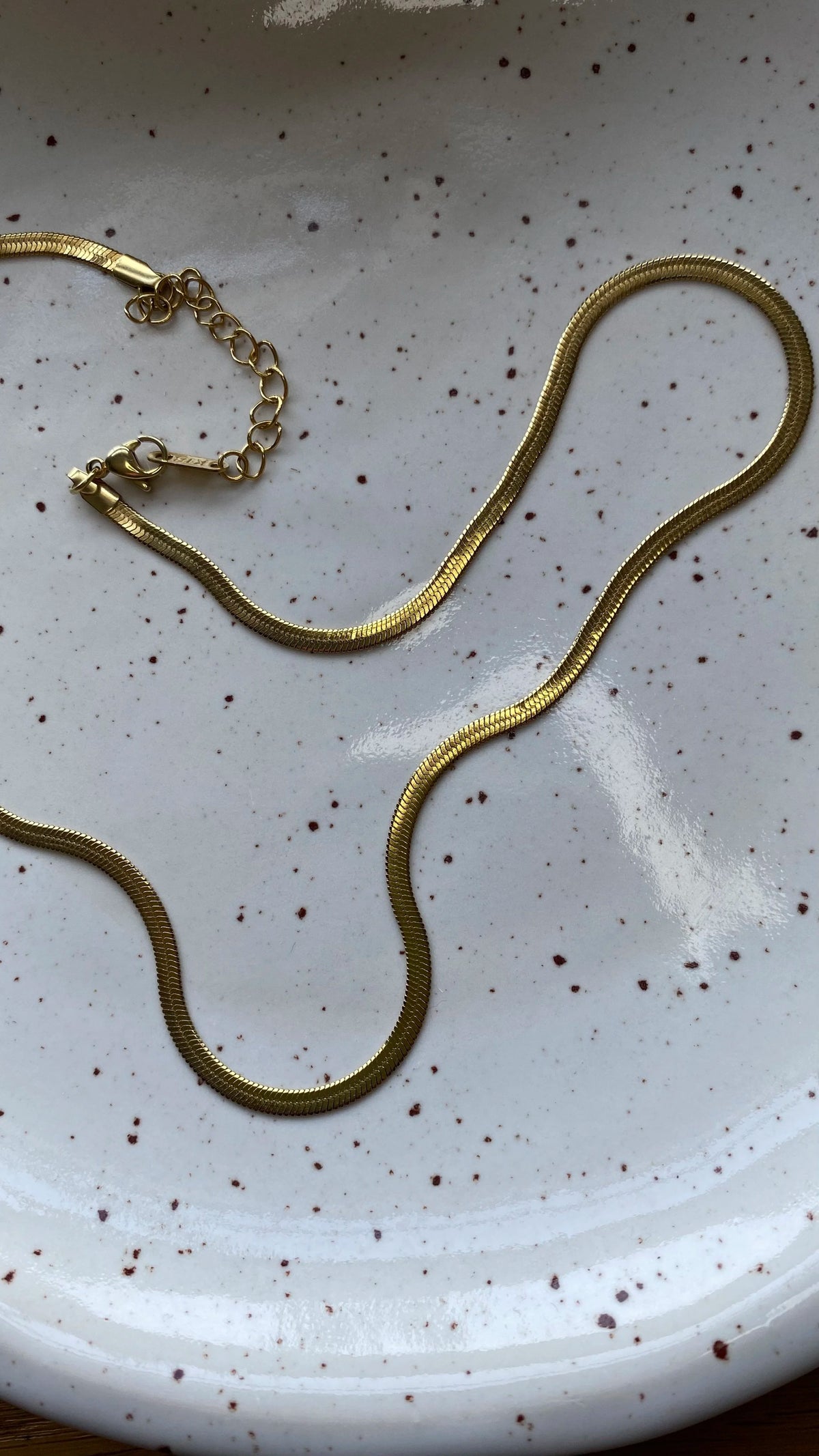 A gold snake chain necklace with a lobster clasp and extension chain, laid out on a white speckled ceramic plate.