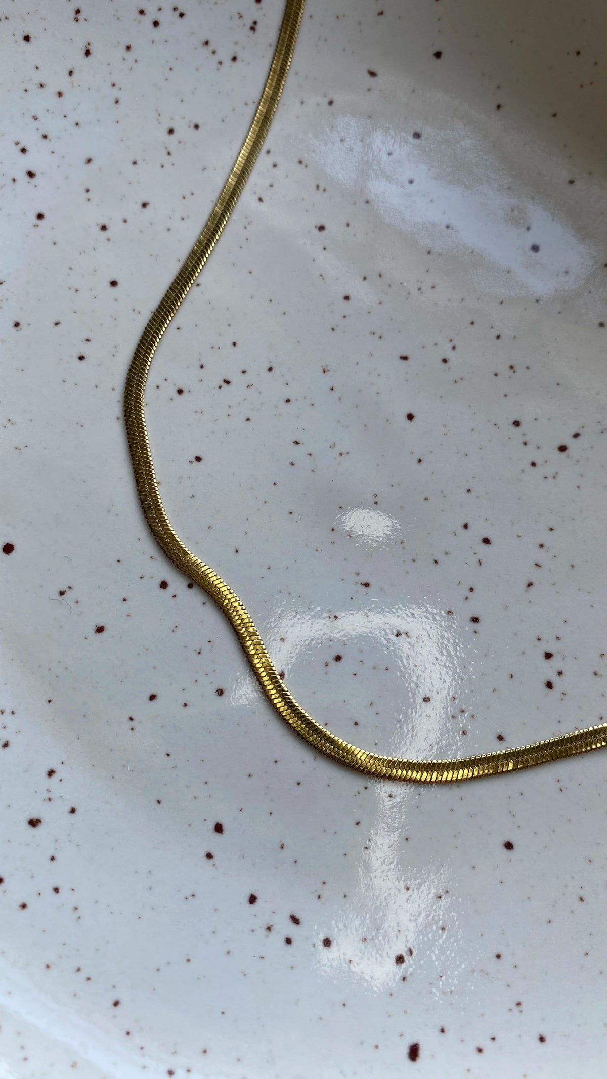A close-up of a gold herringbone snake chain necklace draped over a white speckled ceramic plate.