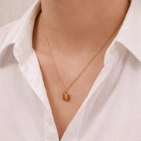 A close-up of a person wearing a delicate gold chain necklace with a small rectangular gold pendant, styled with a white collared shirt.