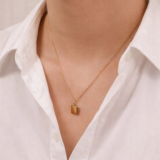 A close-up of a person wearing a delicate gold chain necklace with a small rectangular gold pendant, styled with a white collared shirt.