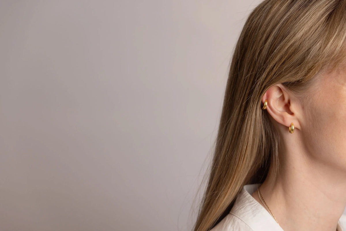 A close-up side profile of a woman with long blonde hair wearing a small gold stack hoop earring in her earlobe and a gold huggie hoop in her ear cartilage, against a plain light grey background.
