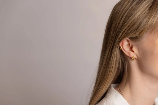 A close-up side profile of a woman with long blonde hair wearing a small gold stack hoop earring in her earlobe and a gold huggie hoop in her ear cartilage, against a plain light grey background.