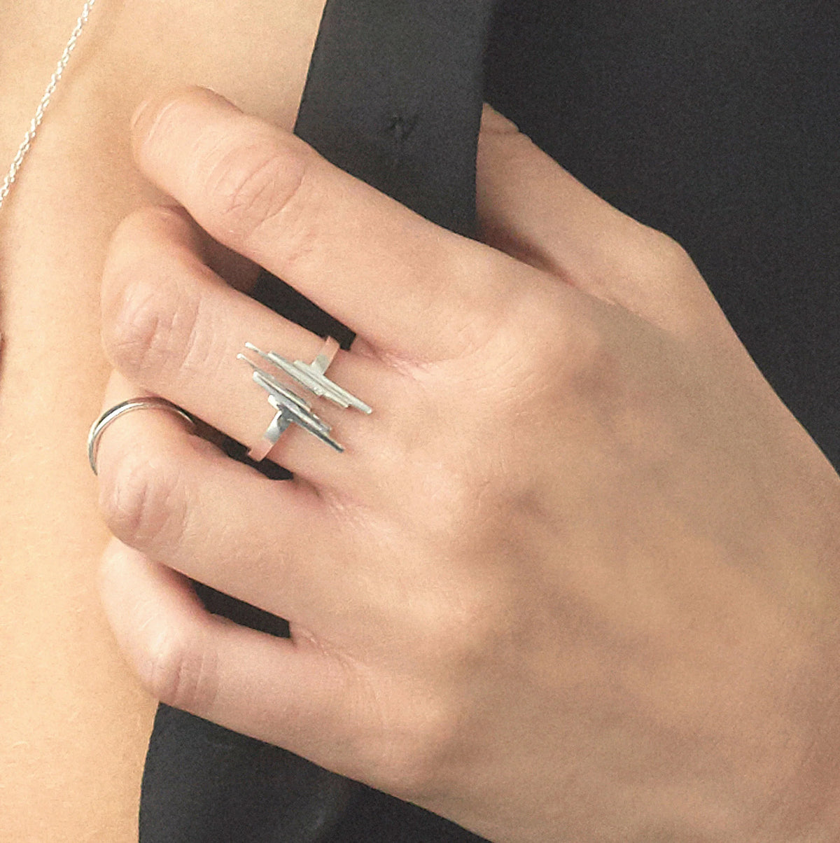A close-up of a hand wearing a silver  Stack ring with three gradating flat bar shapes, alongside a simple silver band on another finger, set against a black top and skin tone.