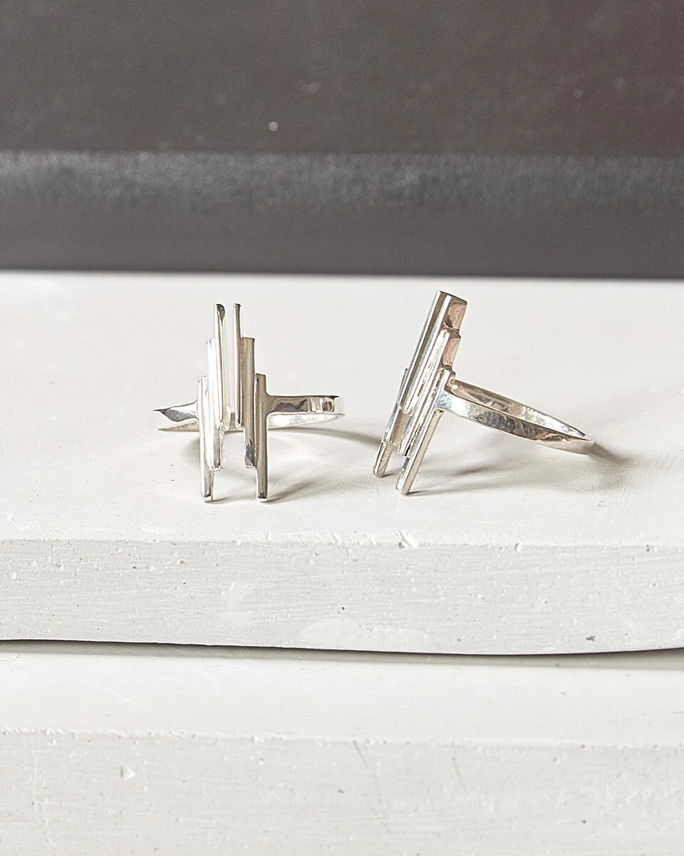 A pair of silver geometric rings with angular, layered bar designs, displayed on a white textured surface with a dark grey background.