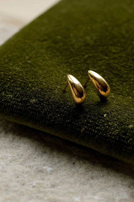 A pair of small gold teardrop-shaped stud earrings resting on a dark green velvet fabric surface.