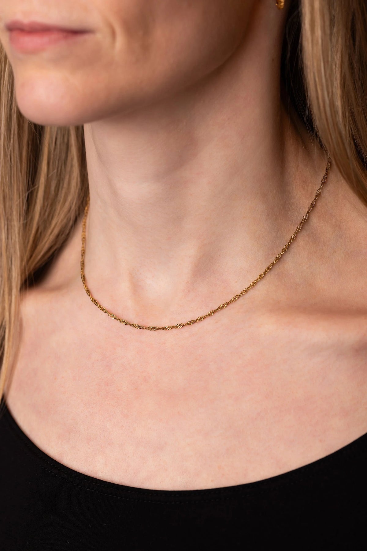 A close-up of a woman's neck and upper chest wearing a delicate gold twist chain necklace and a black top.