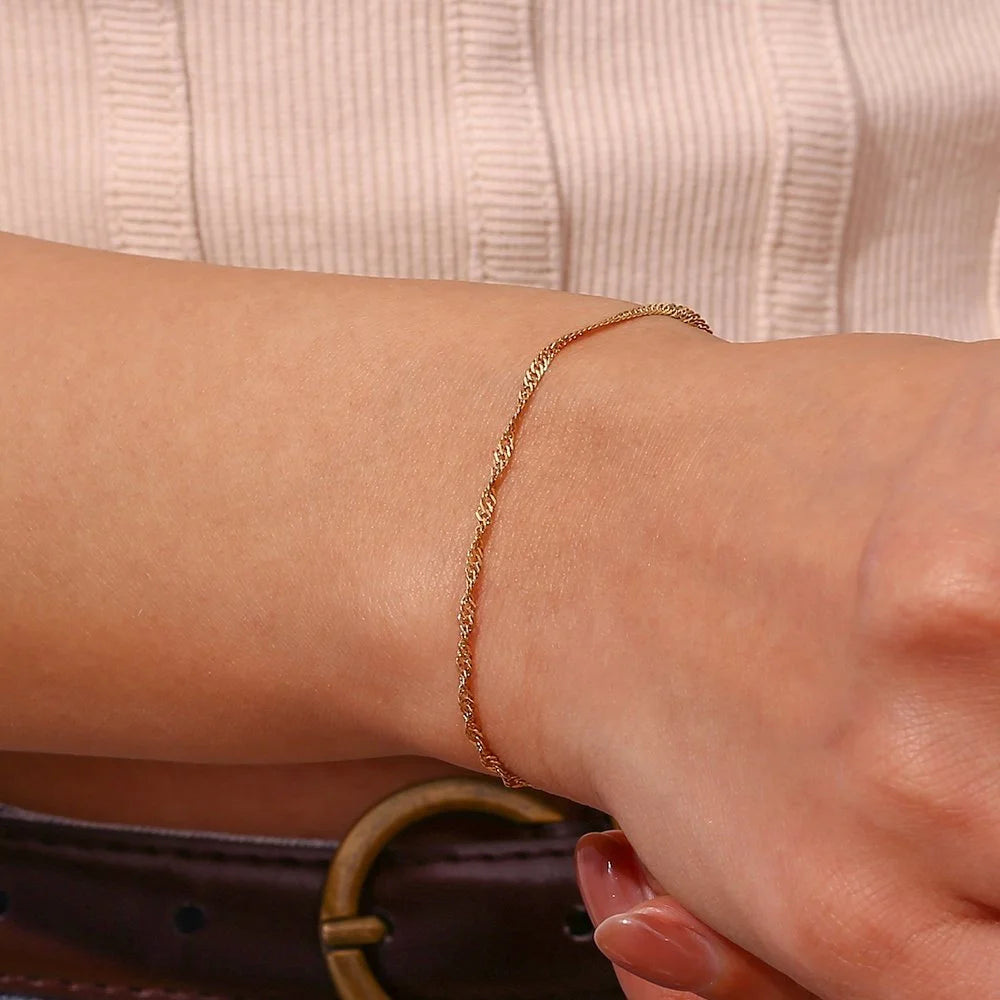 A delicate gold chain bracelet worn on a person's wrist, shown in a close-up lifestyle shot with a ribbed beige top and a dark brown belt visible.