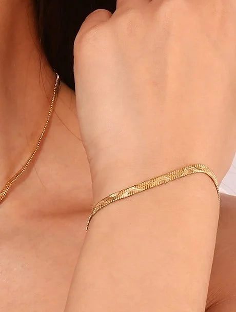A close-up of a person's wrist wearing a gold  Wave chain bracelet, with a matching gold chain necklace partially visible on the shoulder against a light skin-toned background.