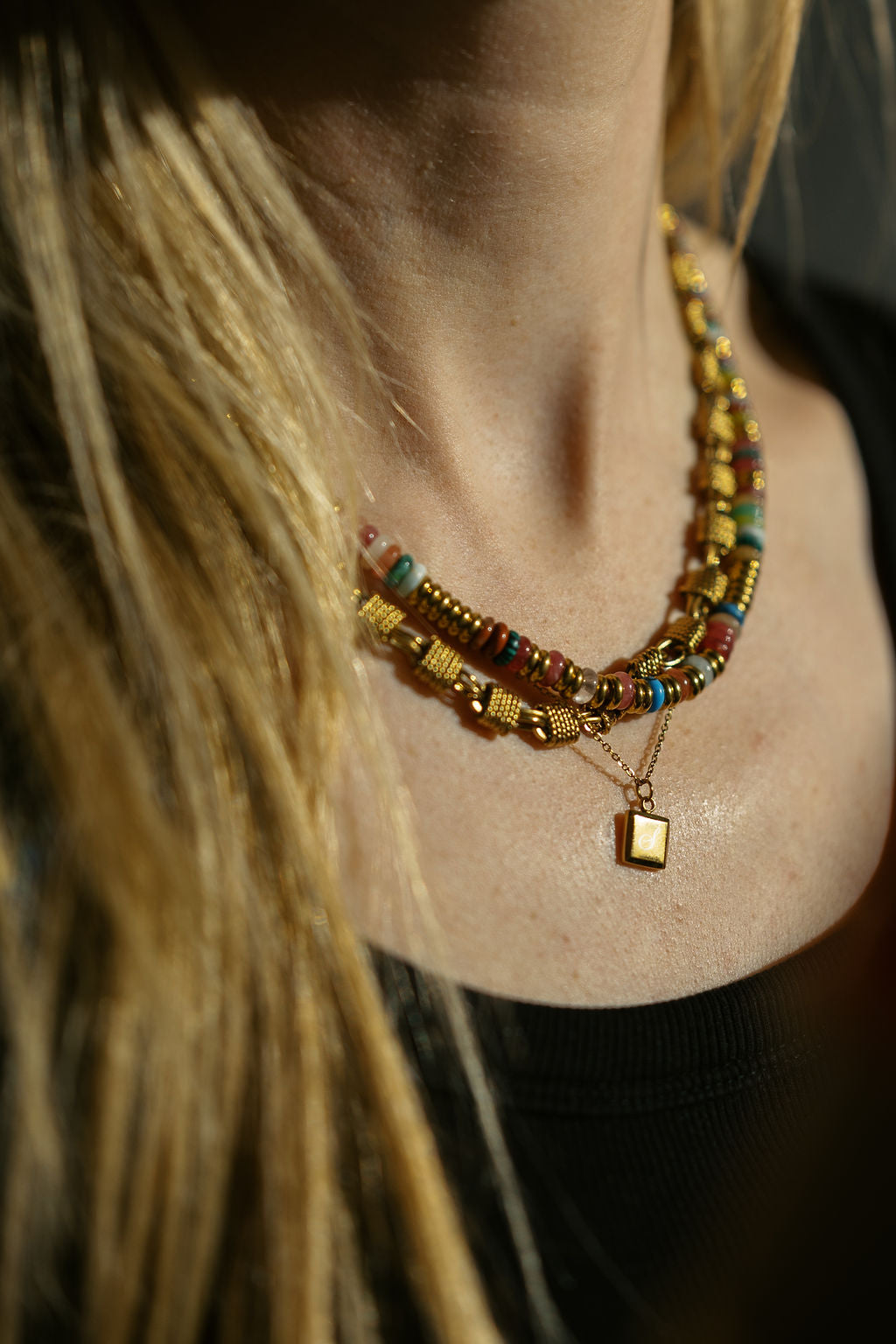 A close-up of a person wearing mixed gemstone necklace with gold-toned beads, turquoise accents, a Woven Chunky chain and a small square frame pendant necklace, against warm golden lighting.