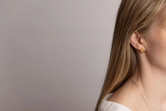 A close-up side profile of a woman with long blonde hair wearing a small gold woven hoop earring and a delicate gold necklace against a plain grey background.