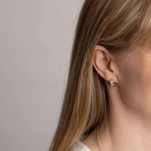 A close-up side profile of a woman with long blonde hair wearing a small silver Woven hoop earring, against a plain light grey background.