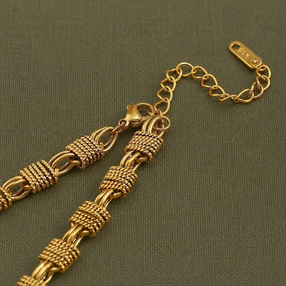 Gold chain necklace with textured woven links. Close up of its adjustable lobster clasp, shown on a dark olive green textured fabric background.