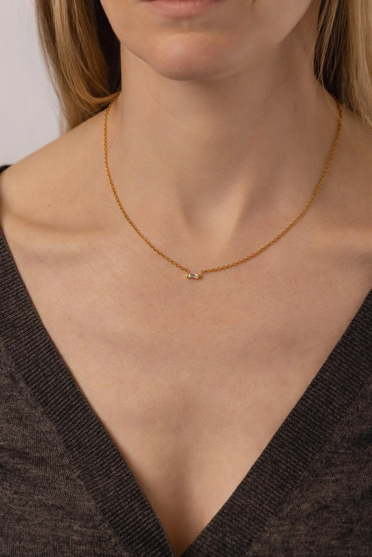 A close-up of a model wearing a delicate gold chain necklace with a small, minimalist gold and cubic zirconia, pendant, paired with a dark gray V-neck top.