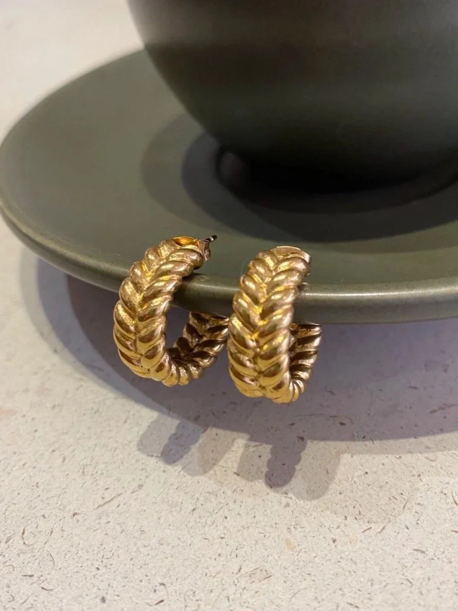 A pair of gold chunky braided hoop earrings resting on the edge of a dark green ceramic saucer, with a neutral light grey surface in the background.
