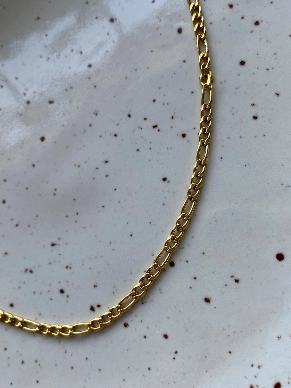 A close-up of a gold Figaro chain necklace resting on a white speckled ceramic plate.