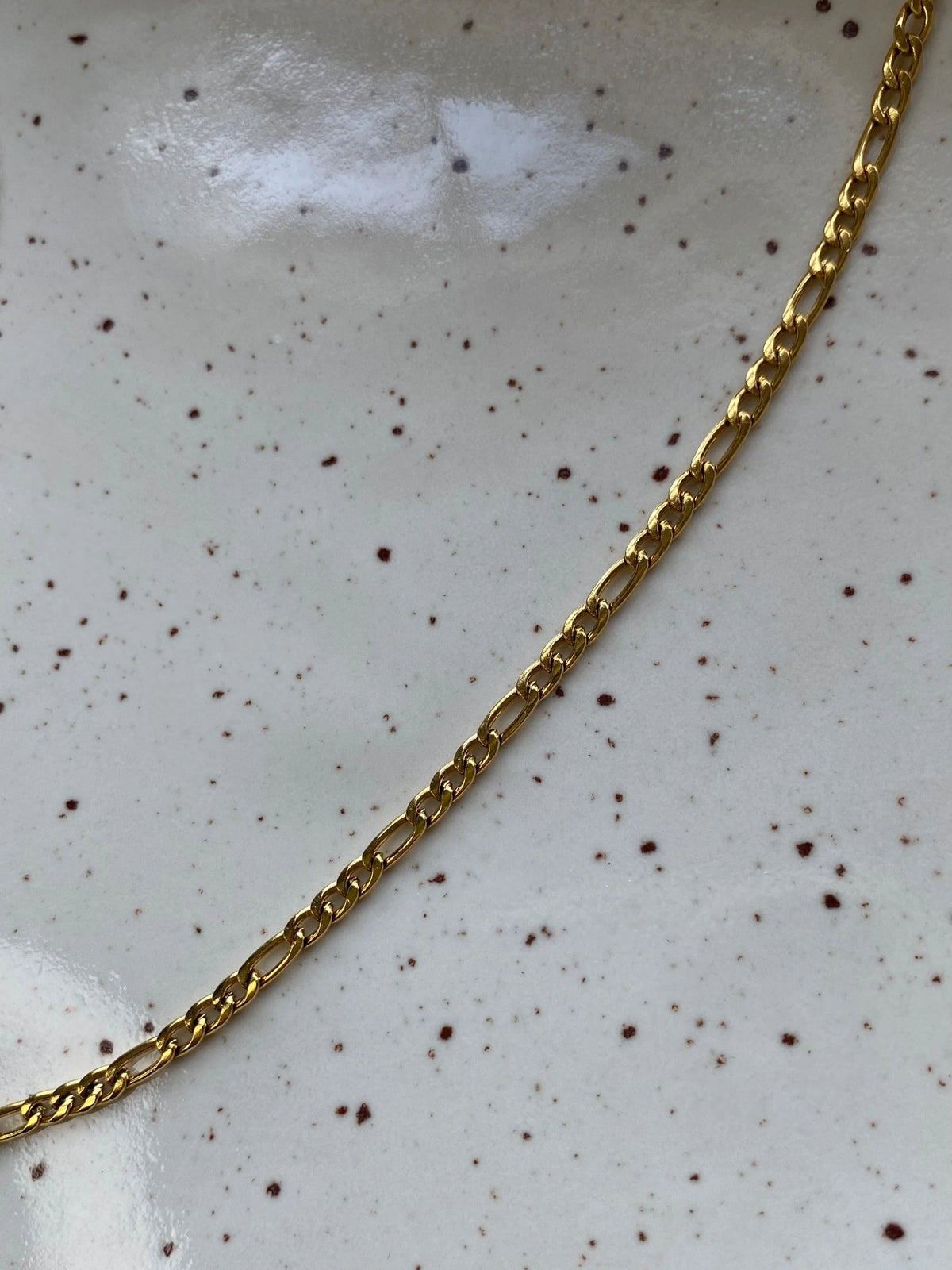 A close-up of a gold Figaro chain necklace laid diagonally across a white speckled ceramic surface.